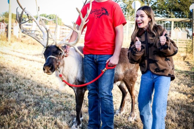 Person smiling beside another holding a reindeer with large antlers on a leash.