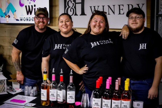 Four people in matching shirts behind a table with J&H Farm & Winery products on display.