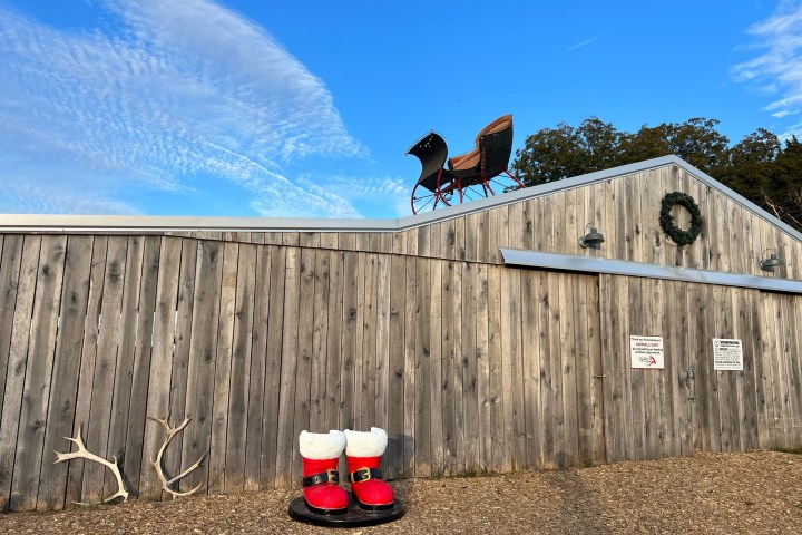 Wooden barn with a sleigh on the roof and large Santa boots on the ground.