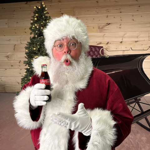 Santa holding a Coca-Cola bottle beside a Christmas tree and sleigh.