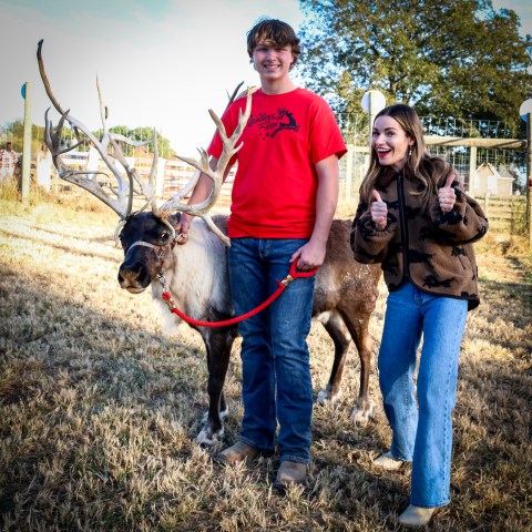 Teen holding reindeer on leash, woman giving thumbs up, outdoors with trees in background.