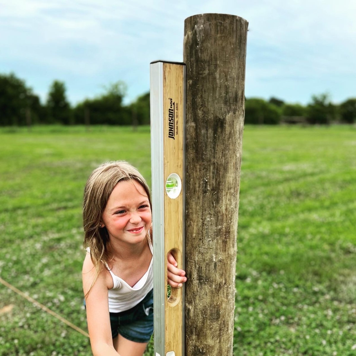 Child smiling, holding a level tool against a wooden post in a grassy field.