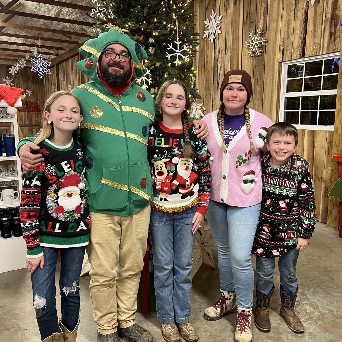 Group of five people wearing Christmas sweaters; standing indoors with festive decorations and a tree.